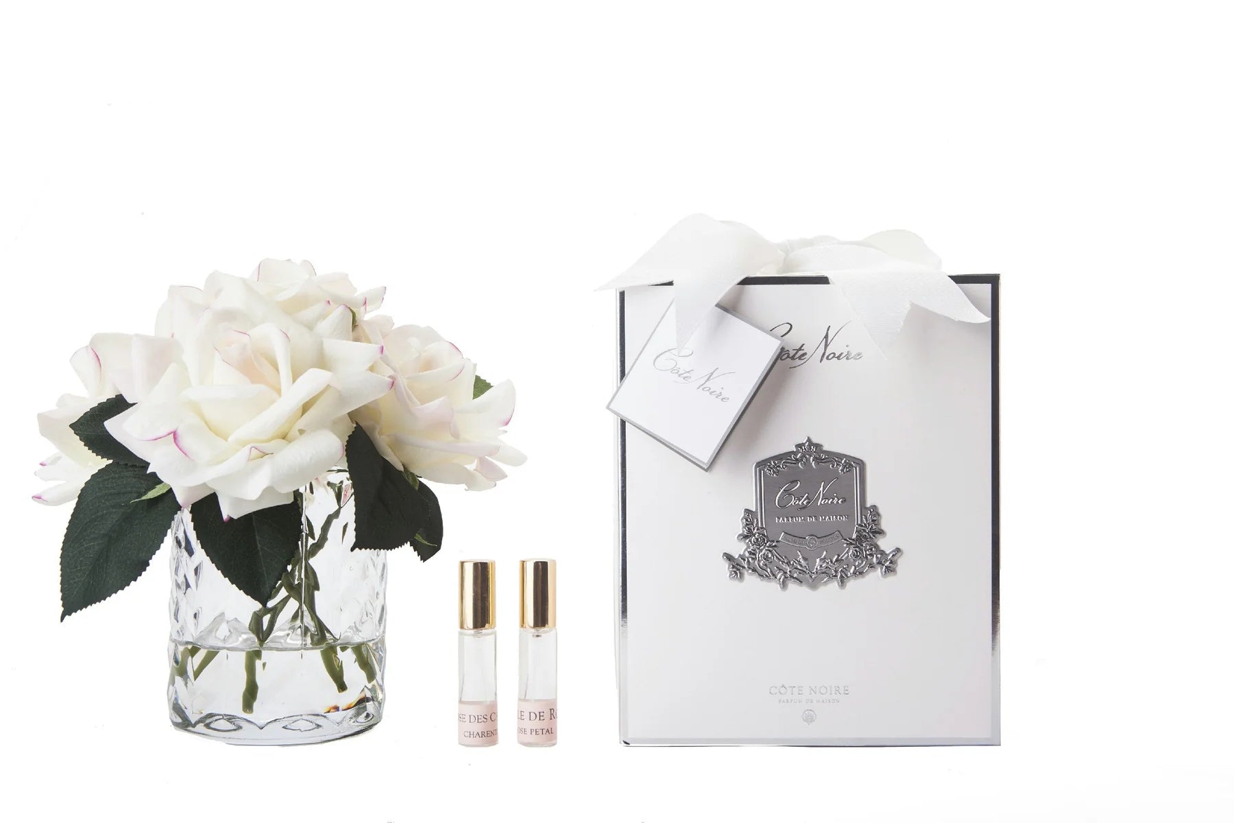 weisse rosen in durchsichtigem glas mit muster. daneben zwei parfumsprays und weisse geschenkbox mit schleife.