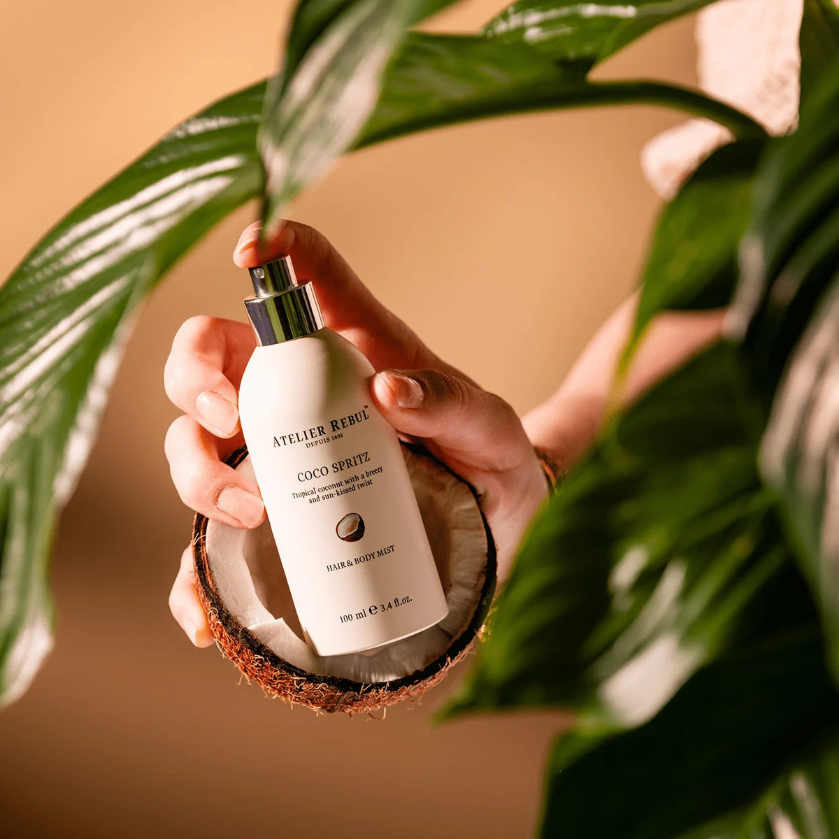 Hände halten das Atelier Rebul Coco Spritz Haar- und Körperspray in einer Kokosnusshälfte und betätigen den Sprühkopf zwischen grünen Blättern.