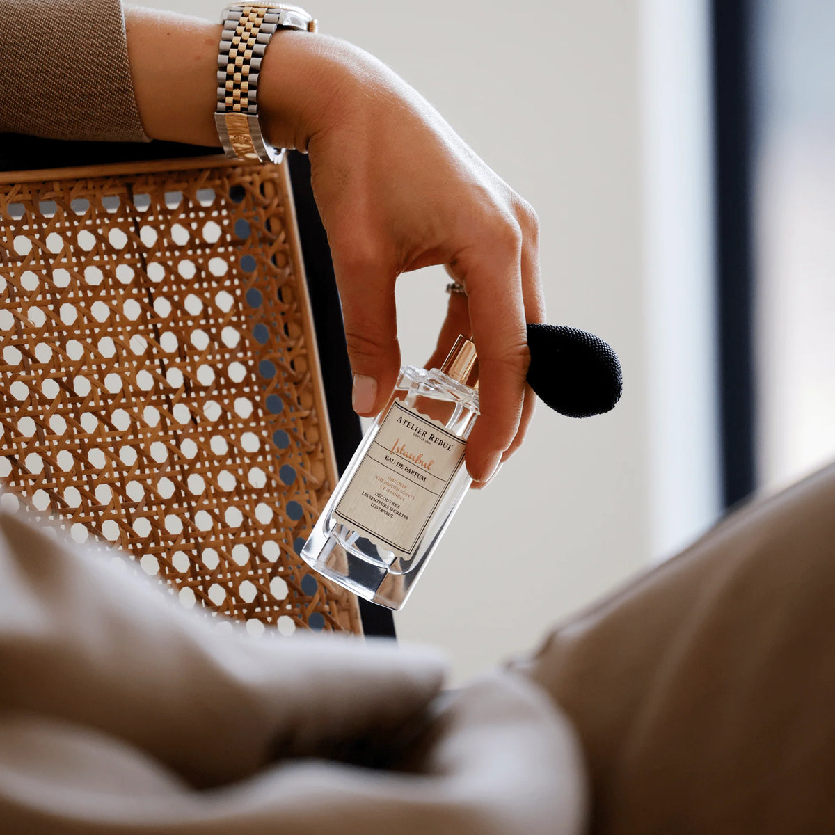 Eine Hand hält Atelier Rebul Istanbul Eau de Parfum 50 ml mit schwarzem Ballonzerstäuber neben einem Stuhl mit Flechtwerk.