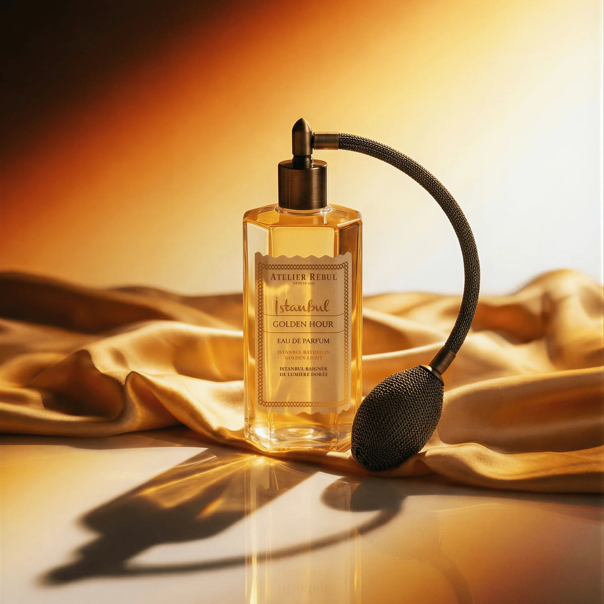 Atelier Rebul Istanbul Golden Hour Eau de Parfum mit Ballzerstäuber, umgeben von glänzendem goldenem Stoff in warmem Licht.