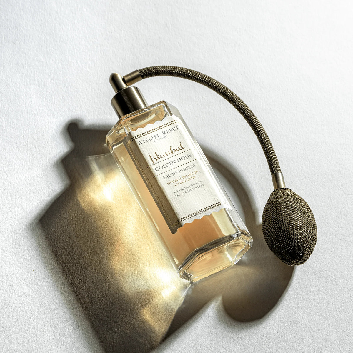 Atelier Rebul Istanbul Golden Hour Eau de Parfum mit olivgrünem Ballzerstäuber, auf hellem Untergrund mit goldenen Lichtreflexen und Schatten.