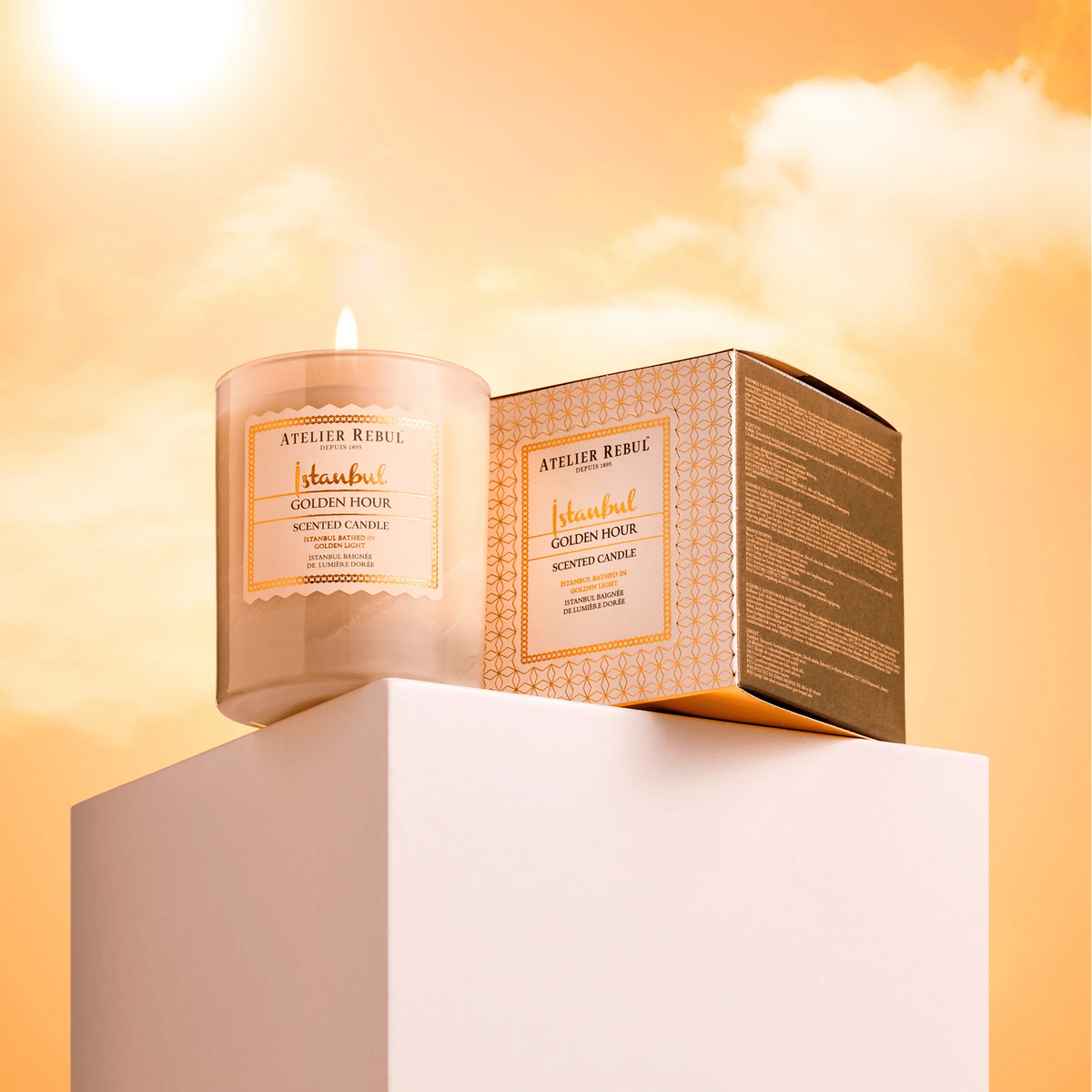 Atelier Rebul Istanbul Golden Hour Duftkerze 210 g mit brennender Flamme und Verpackung auf weißem Podest vor goldenem Himmel.