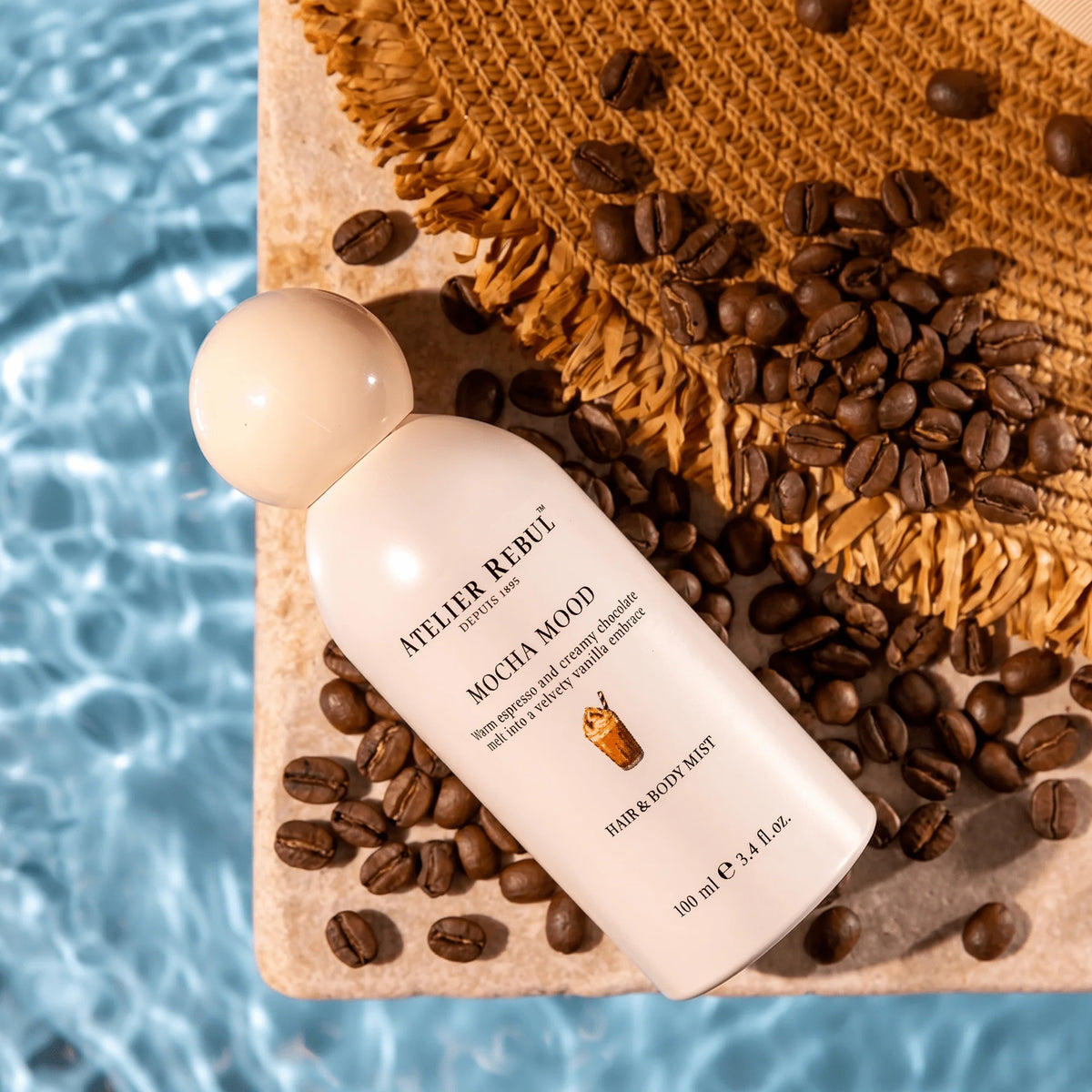 Atelier Rebul Mocha Mood Haar- und Körperspray liegt zwischen Kaffeebohnen neben einer geflochtenen Matte am Poolrand.