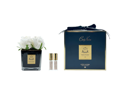 vier weisse rosen in schwarzer quadratischer vase neben zwei pafrumsprays und geschenkbox mit schleife.
