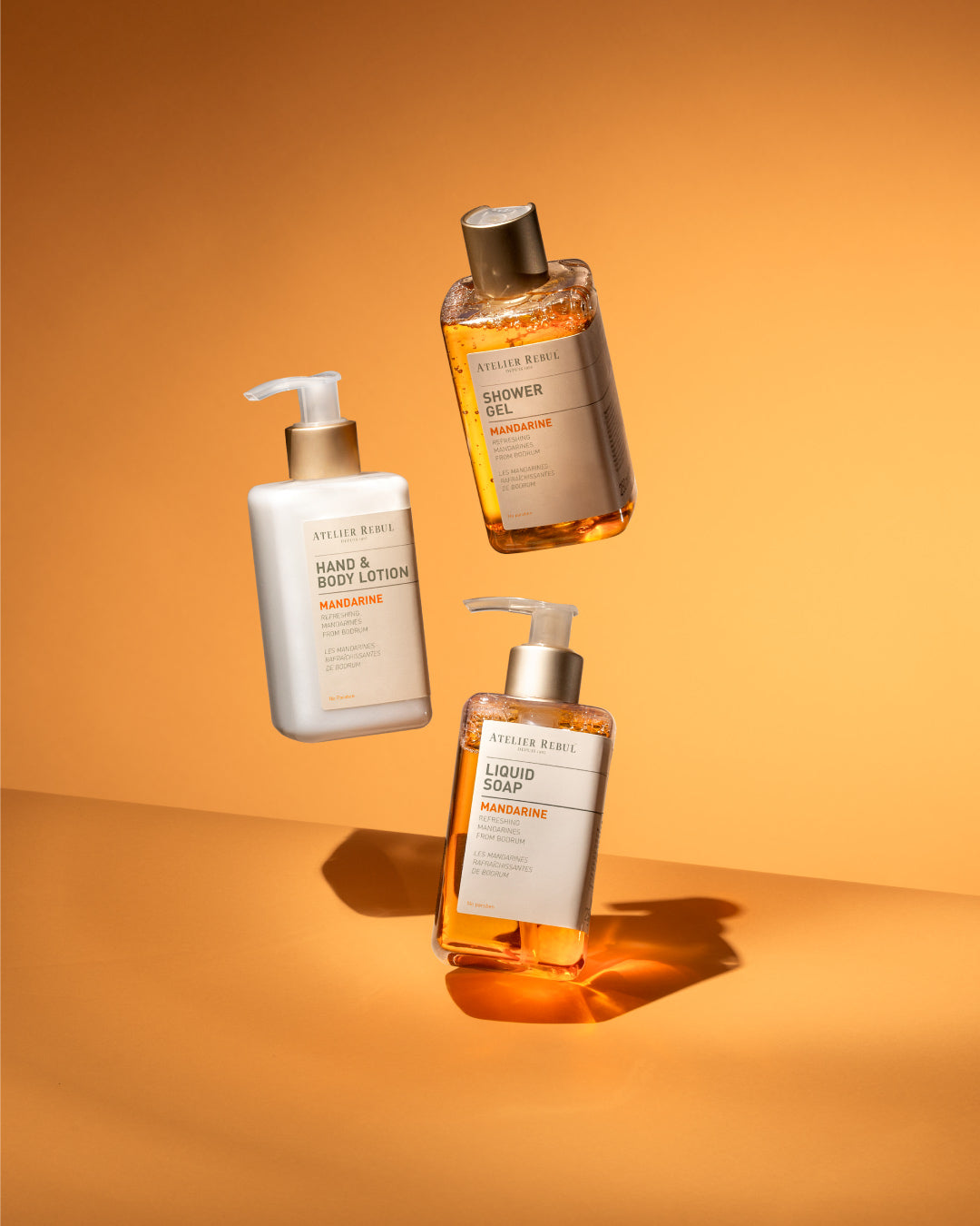 Atelier Rebul Mandarine Hand & Body Lotion, Shower Gel und Liquid Soap, schwebend vor warmem orangenen Hintergrund.