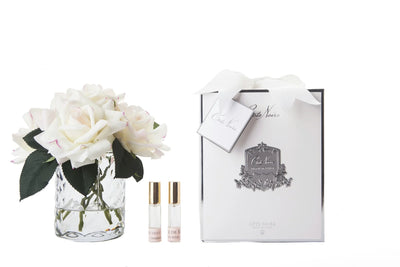weisse rosen in durchsichtigem glas mit muster. daneben zwei parfumsprays und weisse geschenkbox mit schleife.