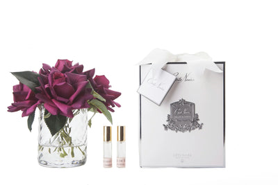 karminrote rosen in durchsichtigem glas mit muster. daneben zwei parfumsprays und weisse geschenkbox mit schleife.