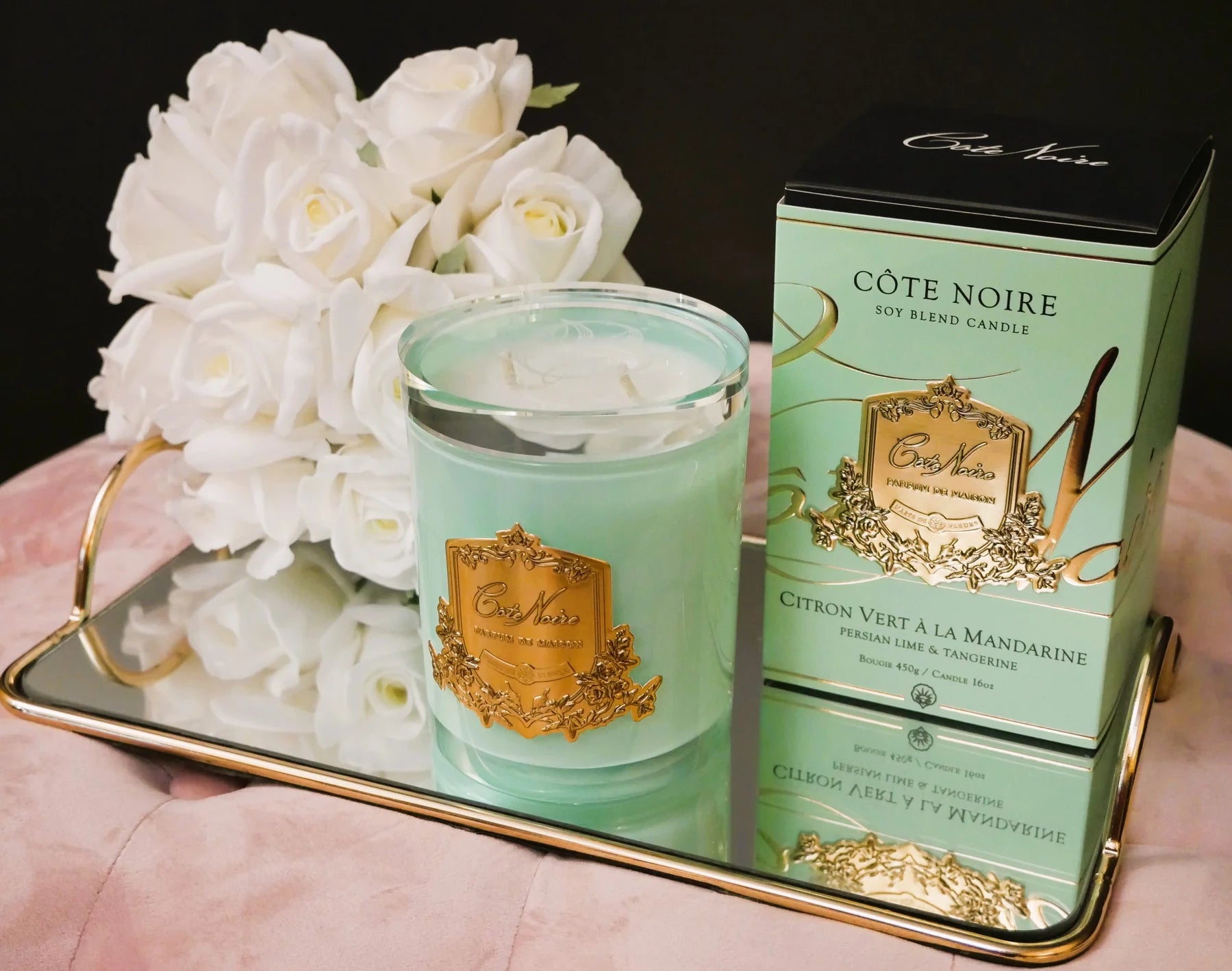 gruene duftkerze mit goldener inschrift, dahinter verpackung und duftblumen in weiss auf goldenem serviertablett. seitenansicht.