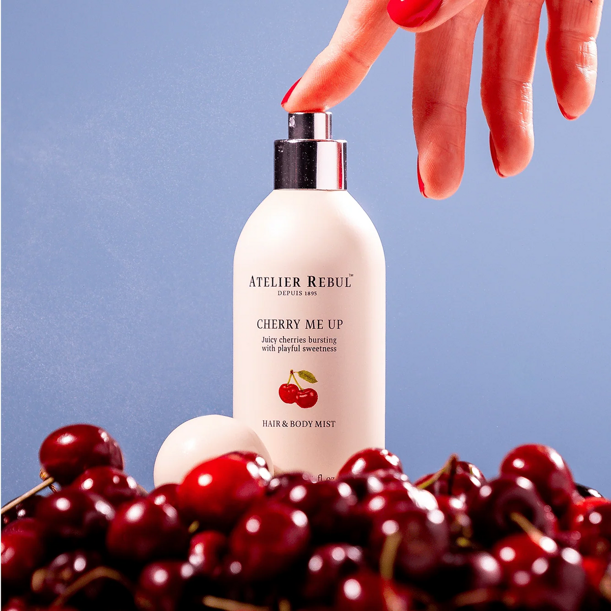 Hand betätigt den Sprühkopf des Atelier Rebul Cherry Me Up Haar- und Körpersprays zwischen roten Kirschen vor blauem Hintergrund.