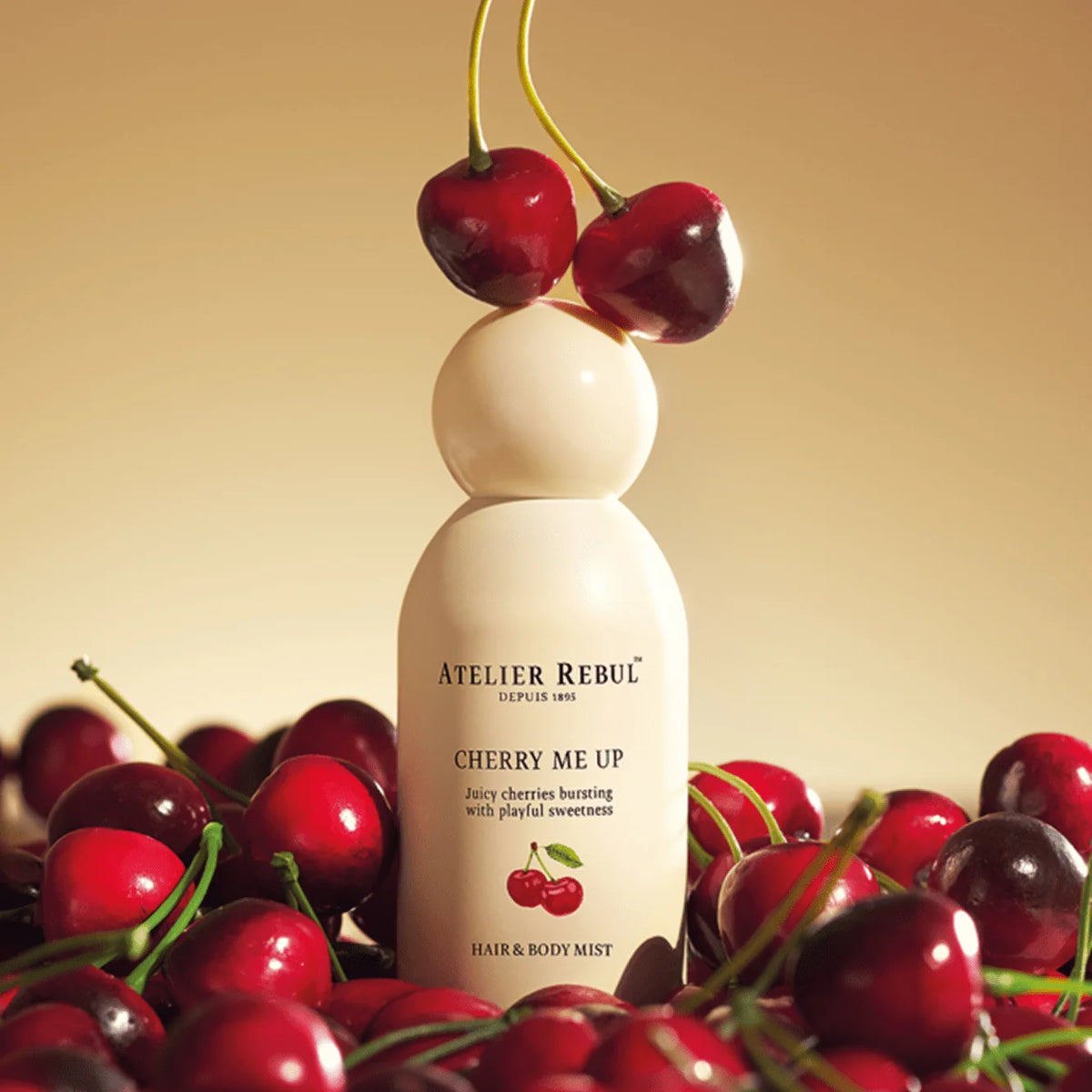 Atelier Rebul Cherry Me Up Haar- und Körperspray zwischen roten Kirschen mit zwei Kirschen auf der runden Verschlusskappe.