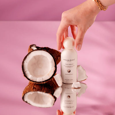 Atelier Rebul Coco Spritz Haar- und Körperspray mit Kokosnusshälften und Kokosstücken auf einer spiegelnden Fläche vor rosa Hintergrund.