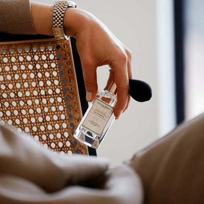 Eine Hand hält Atelier Rebul Istanbul Eau de Parfum 50 ml mit schwarzem Ballonzerstäuber neben einem Stuhl mit Flechtwerk.