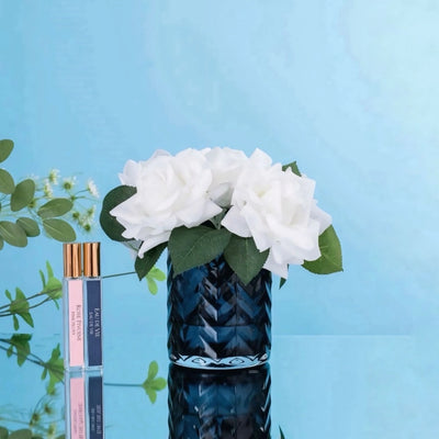 Kuenstliche weiße Rosen in einer blauen Vase neben zwei Parfuemflaschen vor einem hellblauen Hintergrund.