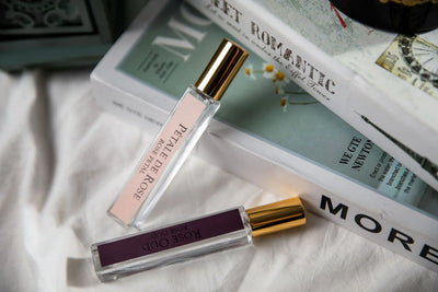 petale de rose duftspray und rose oud duftspray mit goldener kappe vor buechern auf einem weißen tuch.