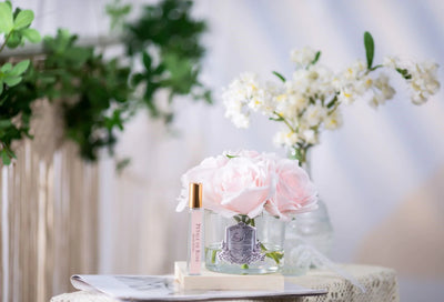 5 pinke duftblumen im glas und duftspray auf weißer unterlage. weitere pflanzen im hintergrund.