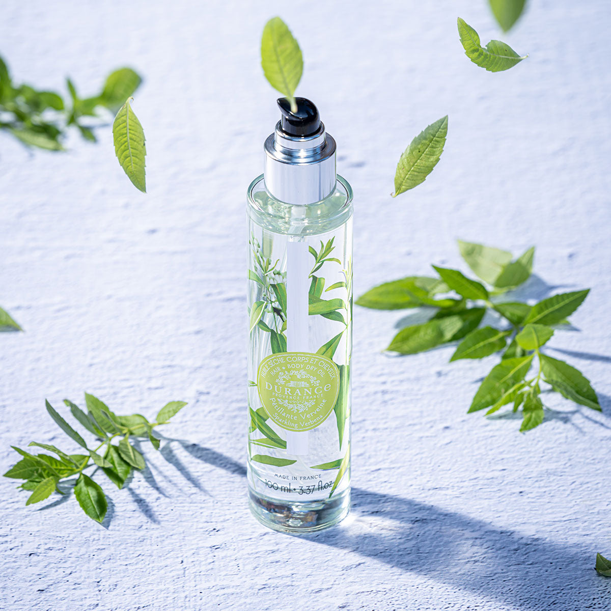 runde hohe glasspruehflasche prickelnde verbene steht auf weissem tisch daneben  blaetter