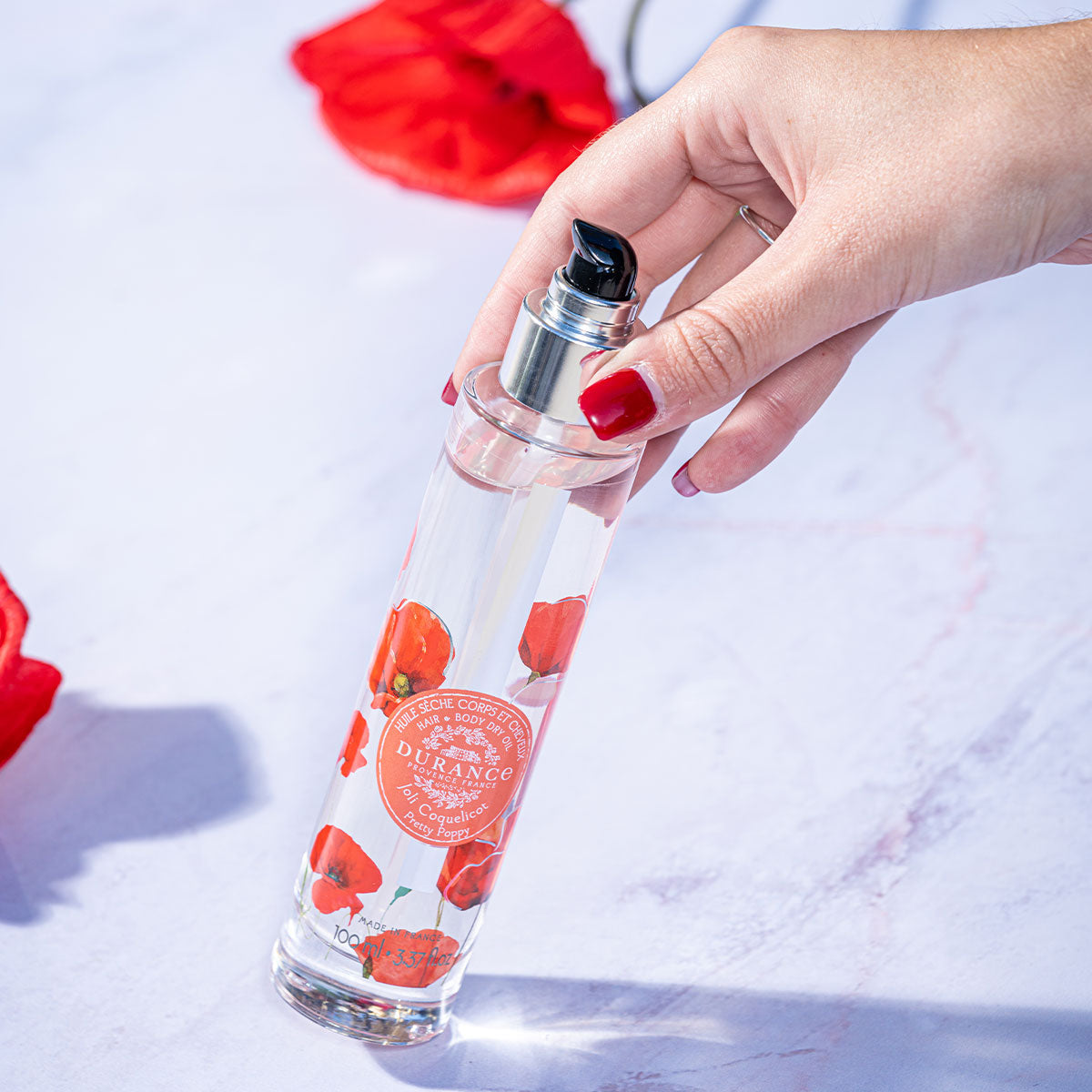 eine hand haelt leichtschraeg die runde glaspumpflasche schoene mohnblume hintergrund weiss mit mohnblumen 